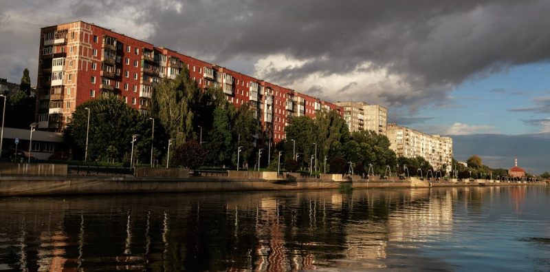 Long Soviet-era apartment buildings line the calm Pregolya River in Kaliningrad under dramatic stormy clouds, with the King's Gate (Korolevskie Vorota) lit up in distance.